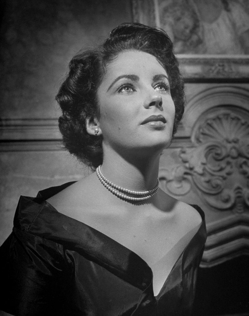 A headshot of Elizabeth Taylor looking upwards with a sleek black off the shoulder dress. Two delicate lines of pearls are draped around her neck and the lighting paints her in a soft glow. 